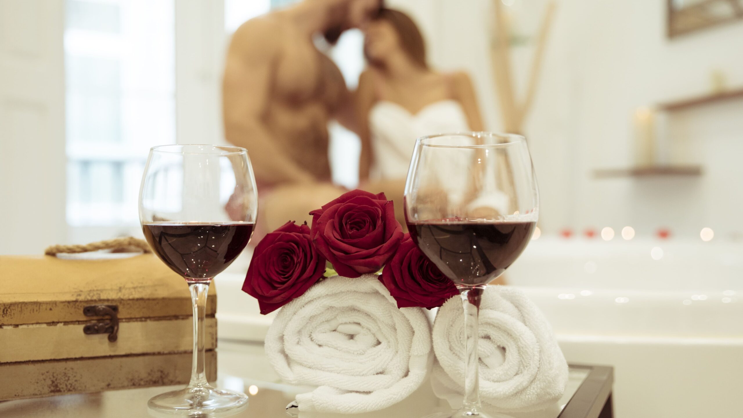 Wine glasses and red roses beside an in-room hot tub with a couple enjoying a romantic stay